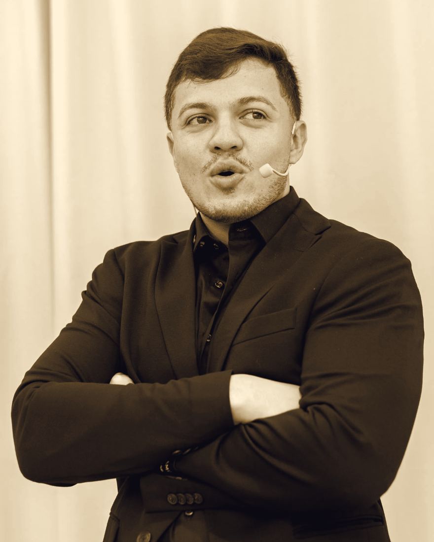 Retrato de Paulo Espíndola de braços cruzados, de blazer preto, com microfone de cabeça